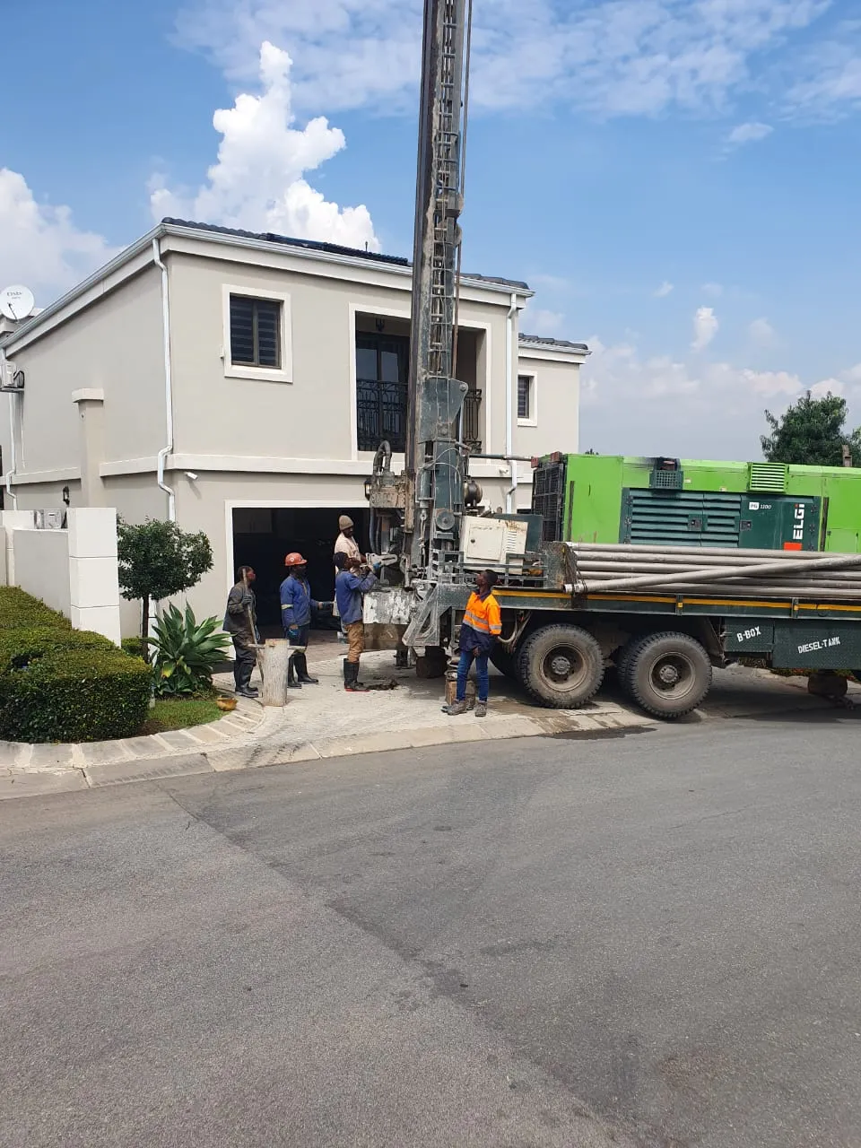 Urgency Boreholes drilling rig and crew drilling a borehole in the driveway of a family home